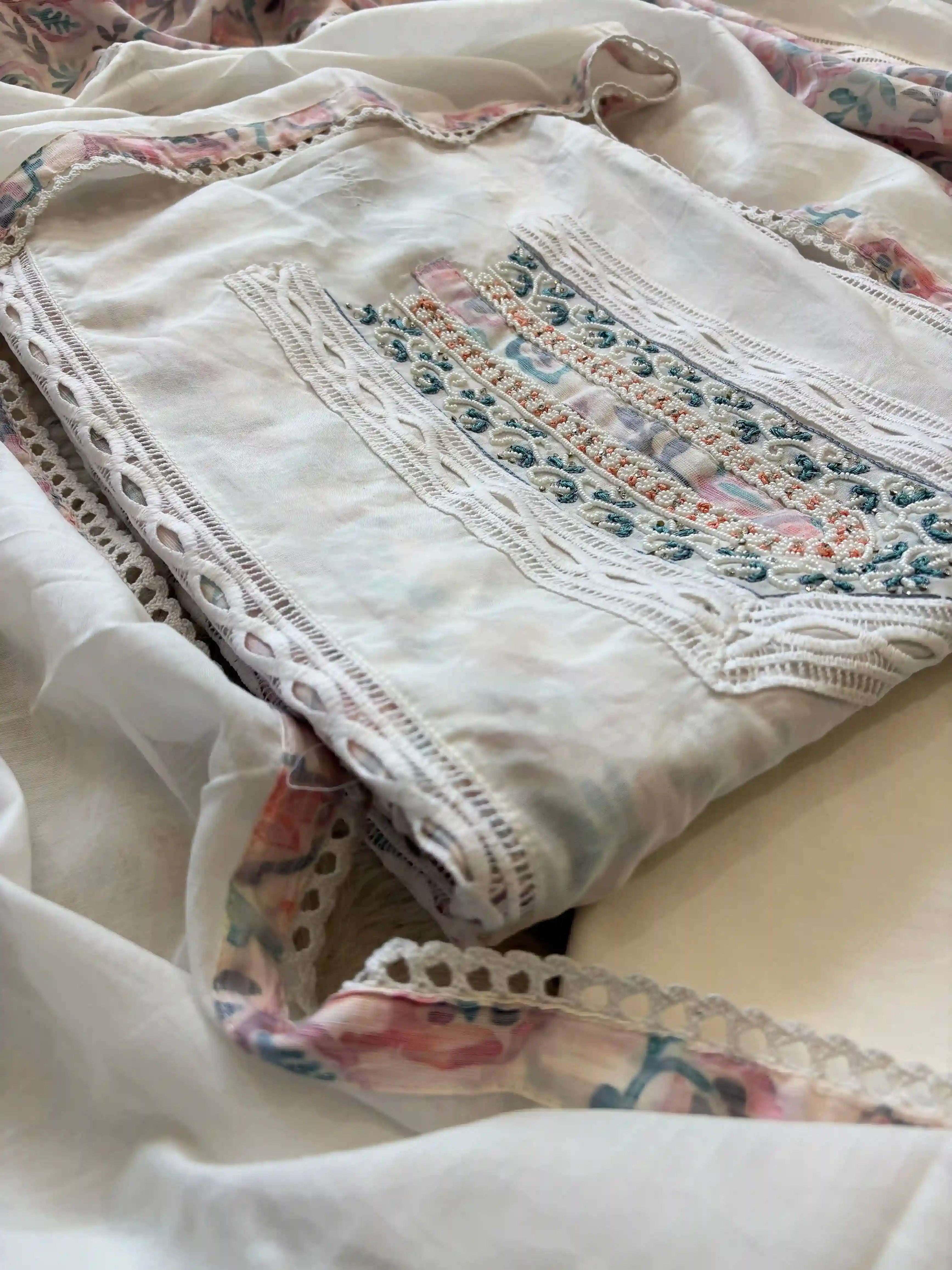 White cotton unstitched suit with embroidered neckline, lace detailing, and pastel floral organza dupatta for summer festive wear.