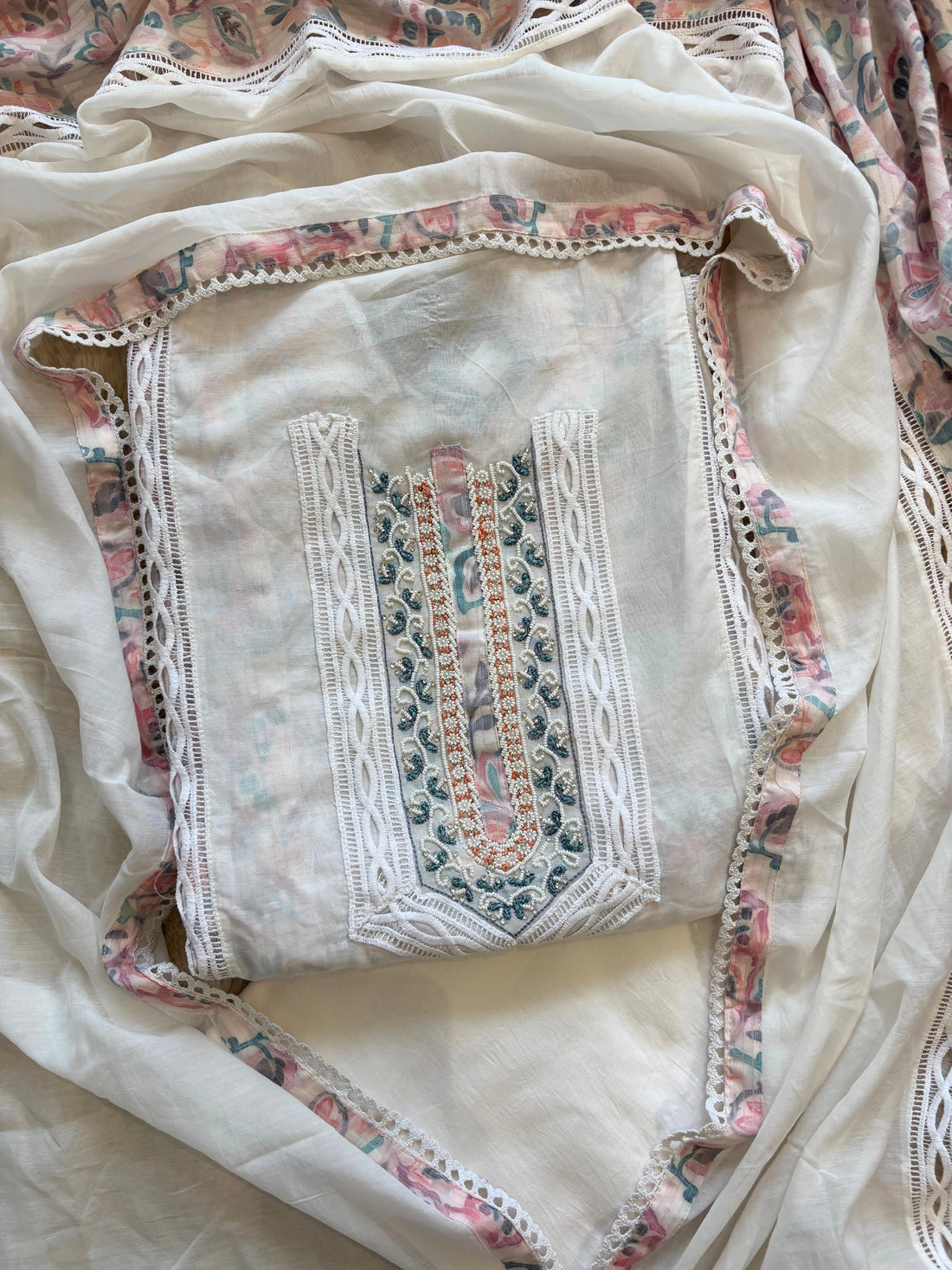 White cotton unstitched suit with embroidered neckline, lace detailing, and pastel floral organza dupatta for summer festive wear.