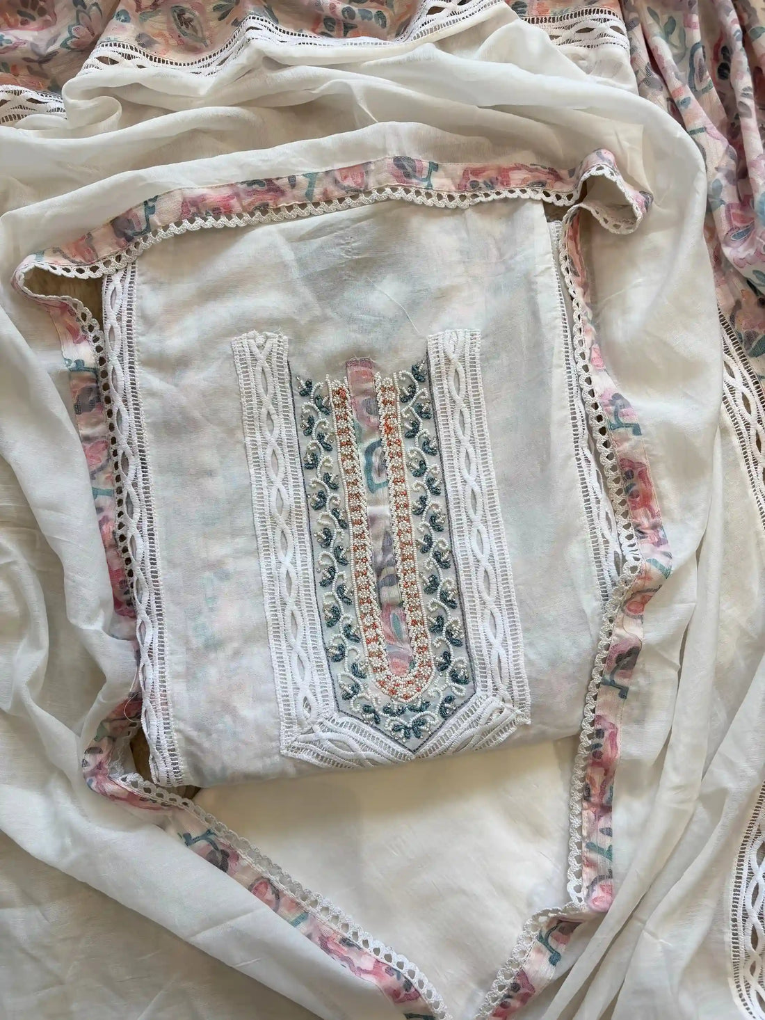 White cotton unstitched suit with embroidered neckline, lace detailing, and pastel floral organza dupatta for summer festive wear.