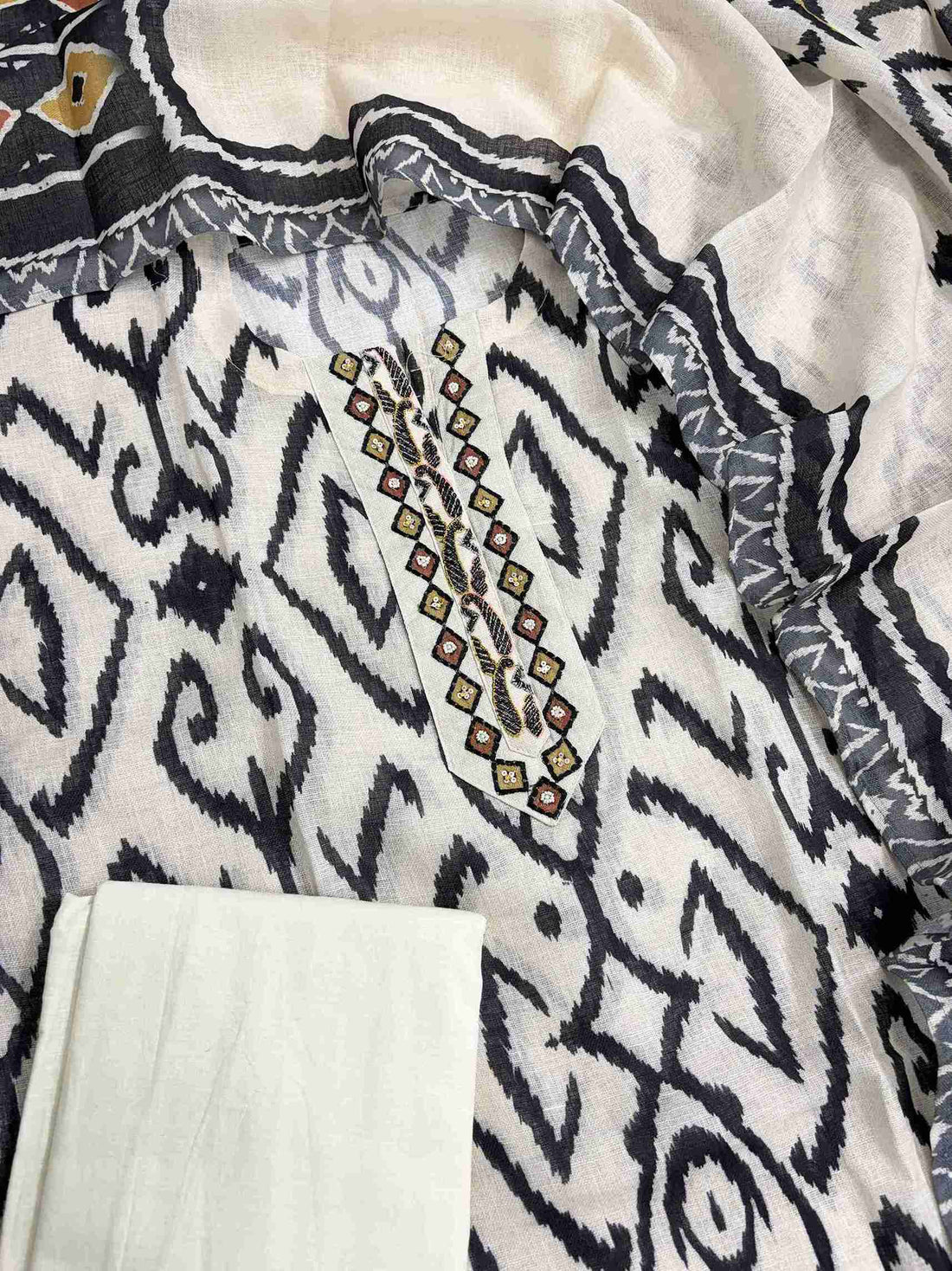 Complete off-white linen unstitched suit flat lay set with black ikat print shirt, ikat border dupatta, and plain cotton bottom